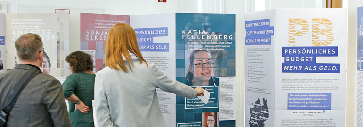 Das Foto zeigt die Wanderausstellung zum Persönlichen Budget und drei Personen, die die Ausstellung betrachten. Die Ausstellung besteht aus vier Elementen. Jedes Element ist ein stehendes Faltdisplay, das Fotos und Texte zeigt. Fünf Personen, die selbst das Persönliche Budget nutzen sind in der Ausstellung porträtiert und ihre Haltung gegenüber dem Persönlichen Budget ist in Zitaten festgehalten. Eine Mitarbeiterin des Landschaftsverbandes Rheinland schildert ihre Erfahrungen mit dem Persönlichen Budget. 