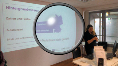 Schulung zur Erstellung barrierefreier Dokumente Ramona Armbrust steht vor einer Wand auf der eine Bildschirmpräsentation ist. Auf der Folie steht "Hintergrundwissen, Zahlen und Fakten". In der Mitte des Bildes ist eine große Lupe eingefügt, die einen Teil des Fotos vergrößert.