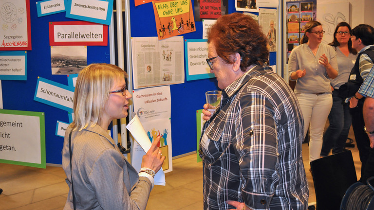 Eindruck von der Veranstaltung. Zwei Frauen unterhalten sich vor einer Pinnwand, auf der Schlagwörter zum Thema Inklusion angebracht sind.