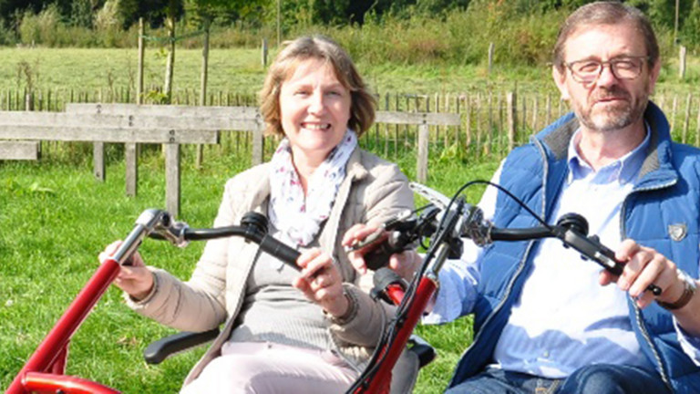 Ein Dreirad-Tandem wird von einem Mann und einer Frau durch eine ländliche und sonnige Umgebung gefahren