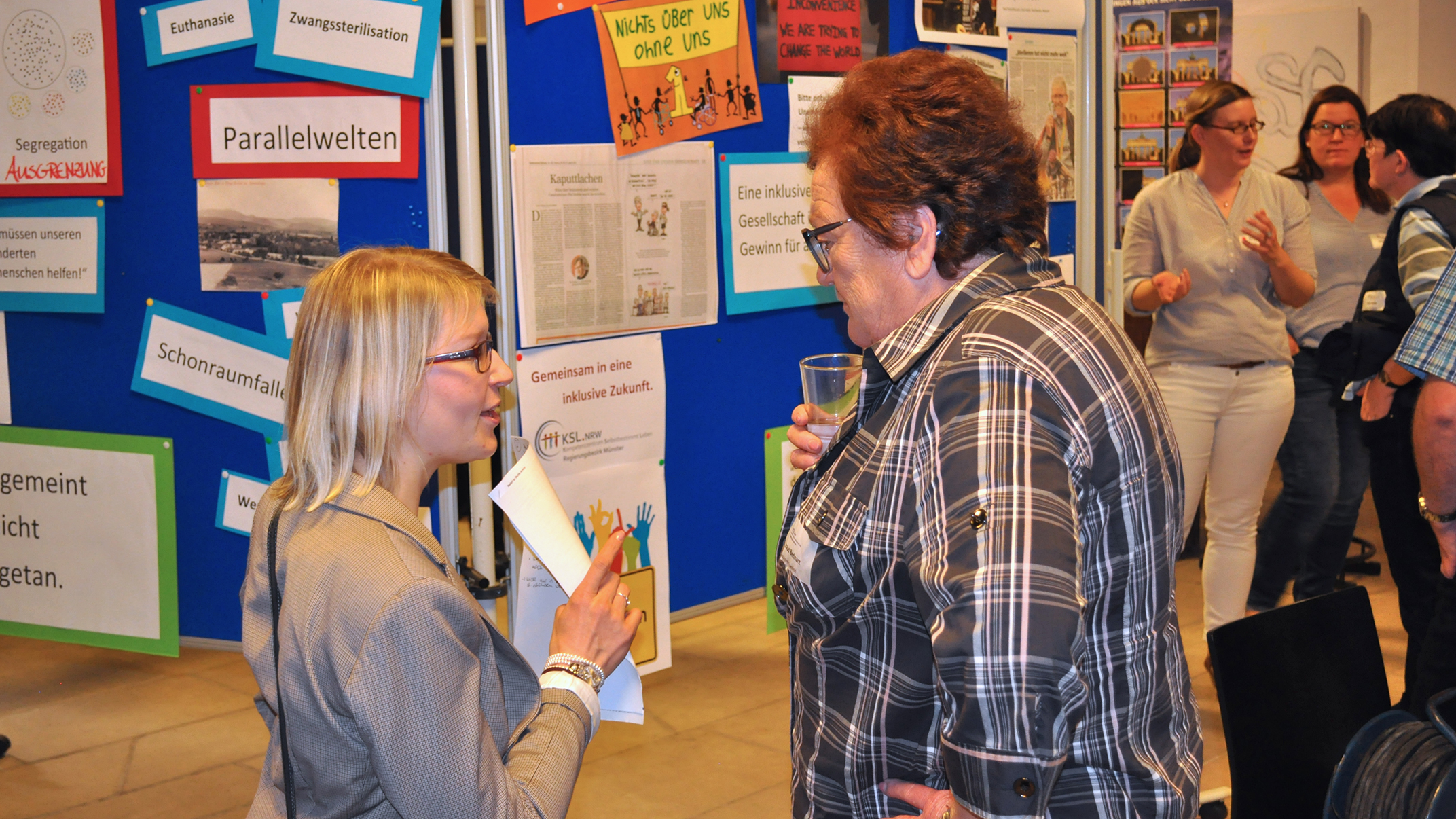 Eindruck von der Veranstaltung. Zwei Frauen unterhalten sich vor einer Pinnwand, auf der Schlagwörter zum Thema Inklusion angebracht sind.