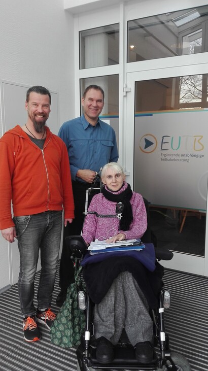 Oliver Schneider vom KSL Münster steht links, in organgener Jacke. Rechts von ihm stehen zwei Mitarbeiter der EUTB Warendorf. Die rechte Mitarbeiterin sitzt im Rollstuhl und trägt einen pinken Pullover. Alle drei stehen vor der Glastür der EUTB Warendorf.